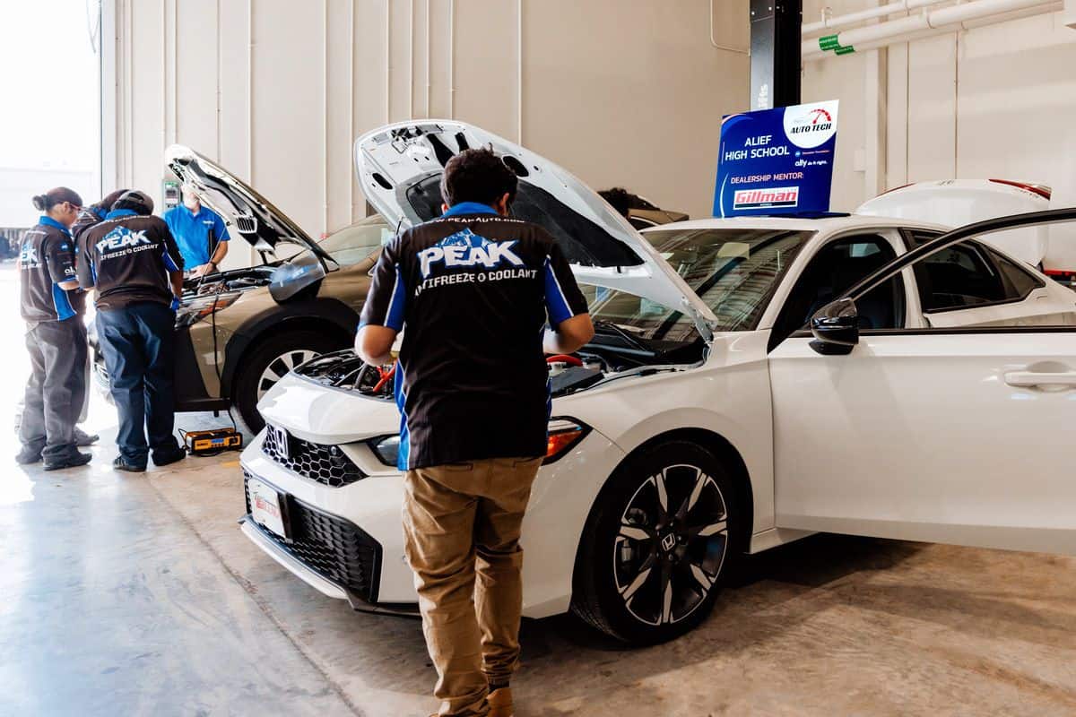 Four students in automotive uniforms work on two cars with open hoods inside a brightly lit auto shop classroom, preparing for an upcoming Auto Tech Competition. Tools and equipment are visible nearby, and a school sign is mounted on the wall in the background.