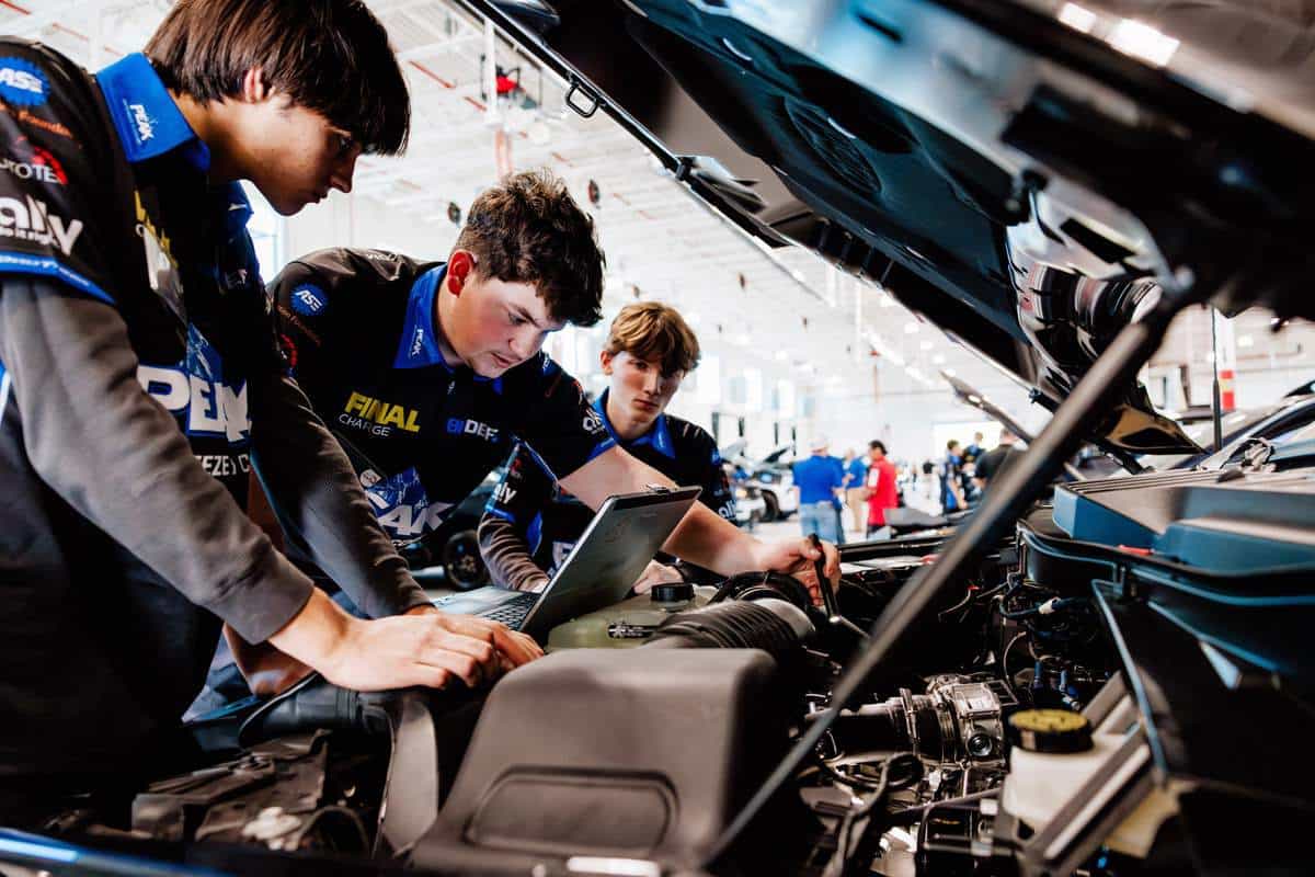 Three young men in racing uniforms examine a car engine while looking at a laptop under the hood in a brightly lit garage, preparing meticulously for the upcoming Auto Tech Competition.