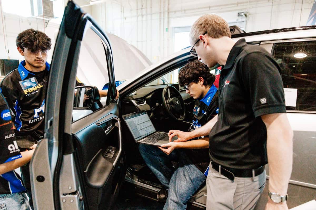 Three people work on a car in an Auto Tech Competition; one sits inside with a laptop, another stands outside looking at the screen, and the third observes. The car door is open in a garage-like setting.