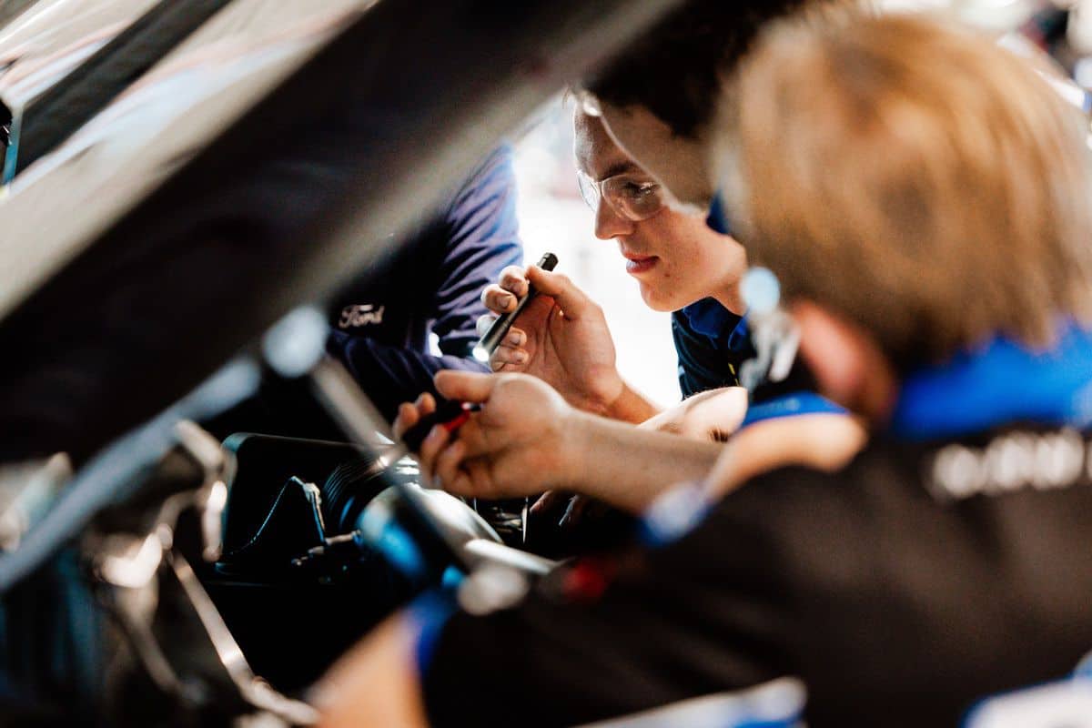 Two people wearing blue work on a car interior, using tools. One person wears safety glasses and focuses on a small part inside the vehicle. The image suggests teamwork and technical focus in an automotive setting, capturing the spirit of an Auto Tech Competition where precision and collaboration are essential.