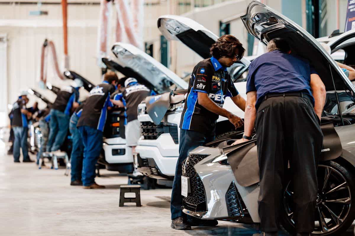Auto technicians work on several cars lined up in a garage, inspecting engines with hoods open. The workers wear uniforms, focusing intently on their tasks in a well-lit, organized workshop, creating an atmosphere reminiscent of an Auto Tech Competition.