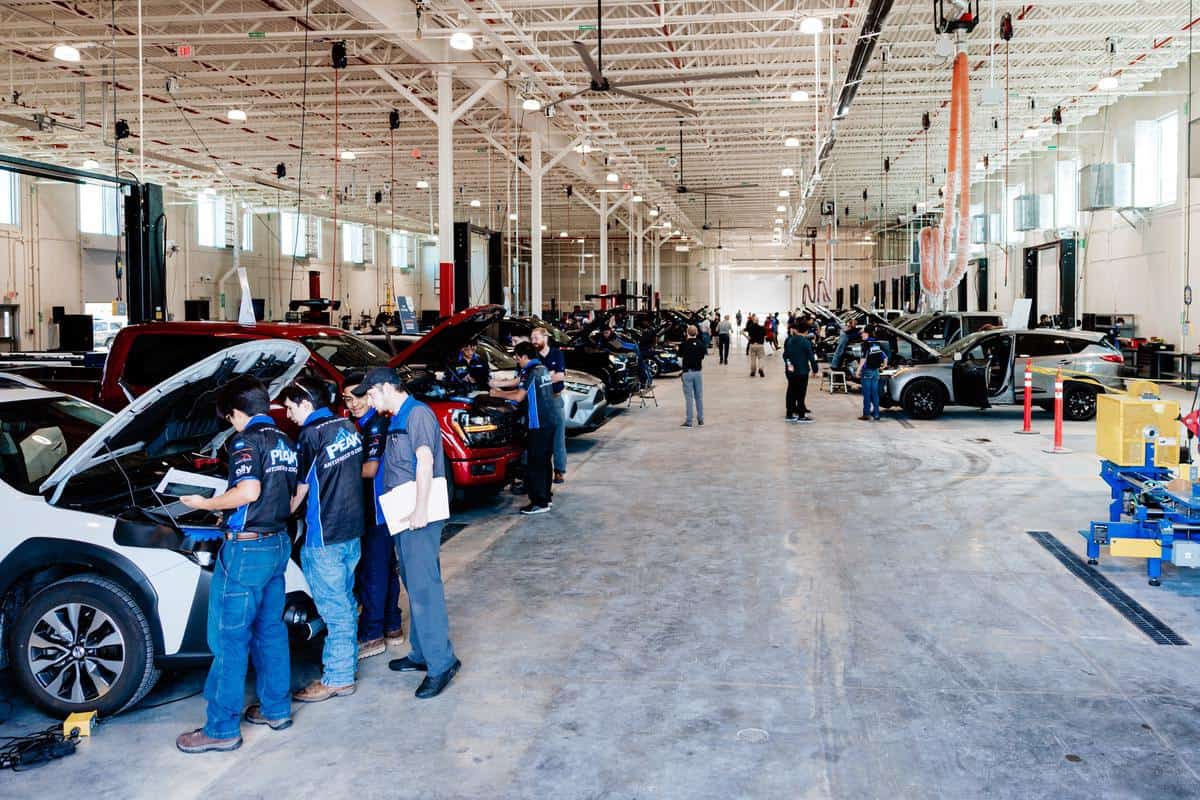 Auto mechanics work on cars in a large, well-lit service garage. Multiple vehicles with open hoods are lined up, while technicians—many of whom are preparing for the upcoming Auto Tech Competition—inspect and repair them. The space is spacious and organized, with equipment and tools visible.