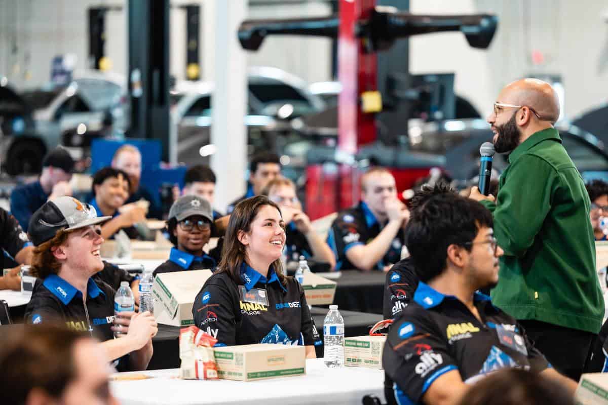A group of people sit at tables with pizza boxes, listening to a speaker holding a microphone. Some attendees are smiling and wearing matching dark uniforms and caps, suggesting their participation in the Auto Tech Competition. The setting appears to be a lively team event.