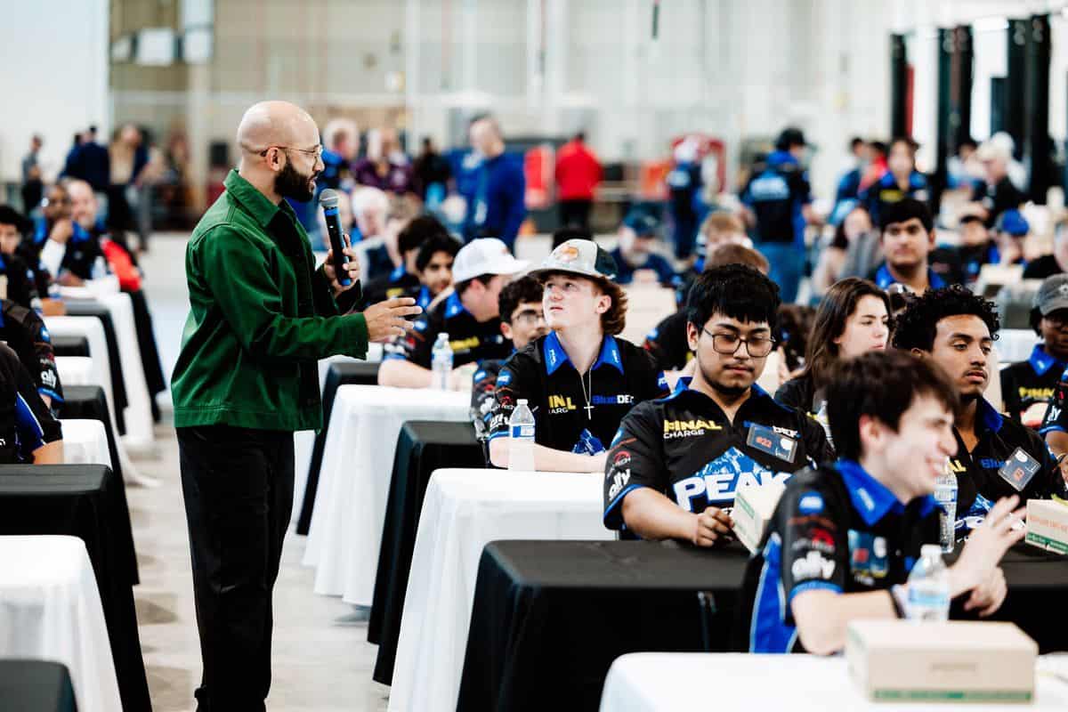 A man in a green jacket speaks to a group of young people seated at tables, possibly during an Auto Tech Competition or workshop, in a large, bright indoor space.