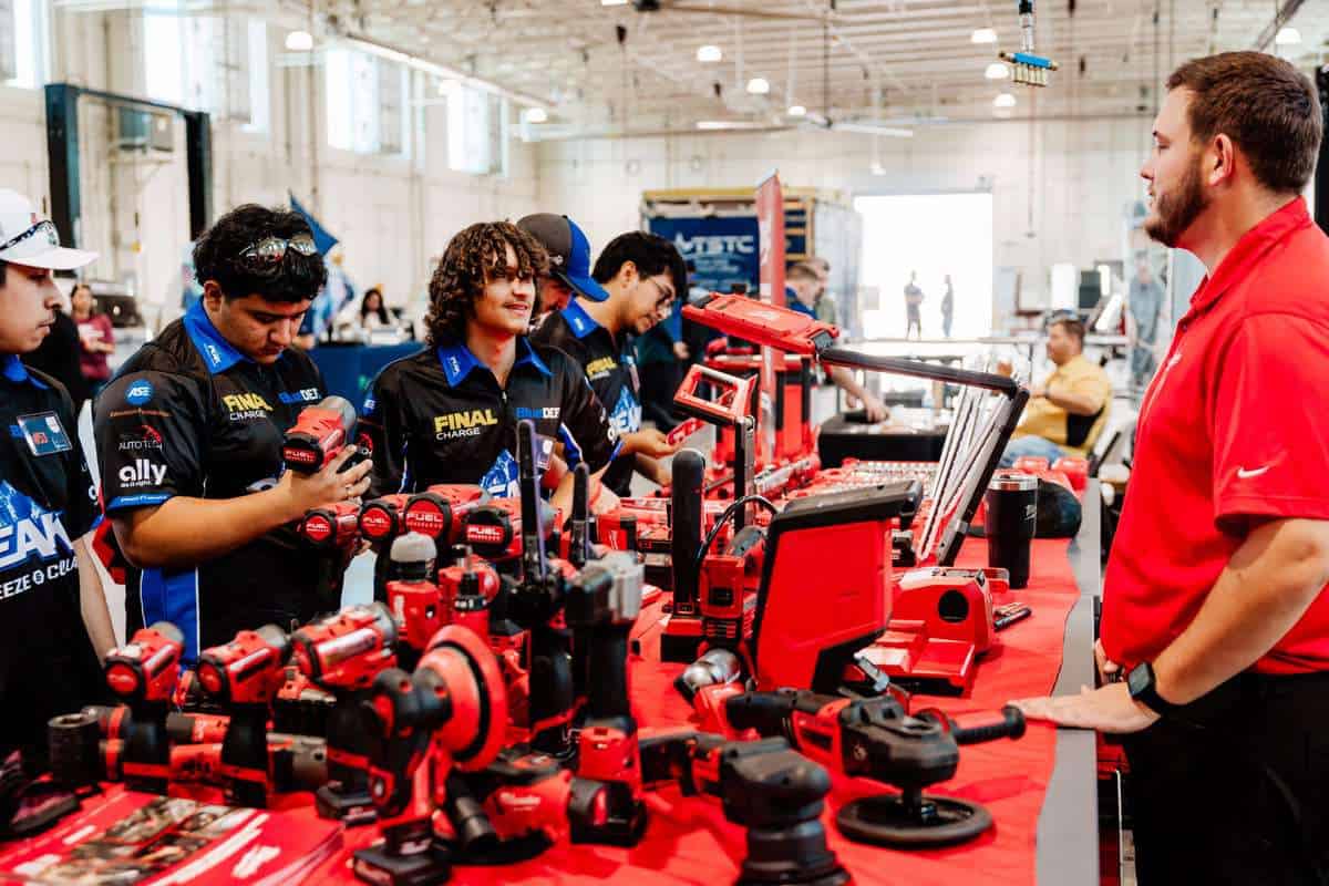 A group of young people look at red power tools displayed on a table, while a man in a red shirt speaks to them inside a well-lit workshop during an Auto Tech Competition.