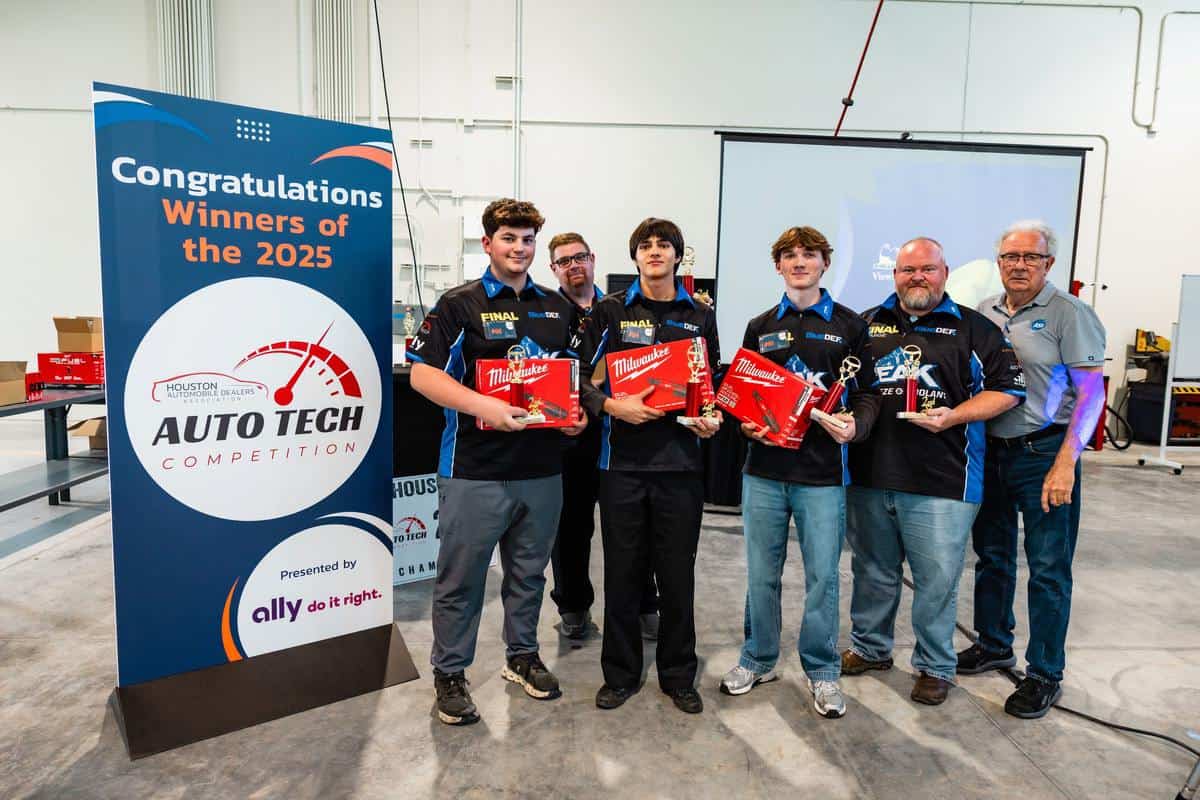 Five men stand together holding prizes next to a sign that reads “Congratulations Winners of the 2025 Auto Tech Competition.” The group appears to be celebrating in an industrial indoor space.