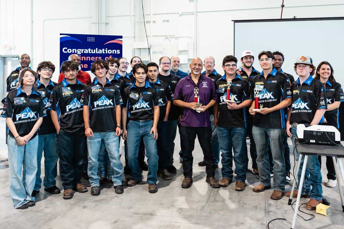 A group of students and adults stand indoors, many wearing matching "PEAK" shirts, celebrating their success in the Auto Tech Competition. Two people hold trophies as a "Congratulations Winners" sign is visible in the background. Everyone is smiling.