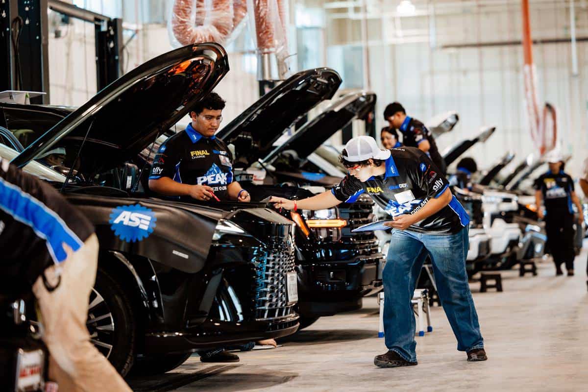 Automotive students in matching uniforms inspect cars with open hoods inside a large, well-lit garage, using clipboards and diagnostic tools during an Auto Tech Competition or workshop.