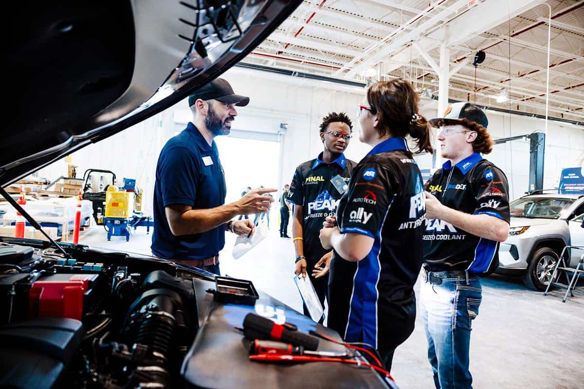 A man in a cap speaks to three people in racing uniforms inside a garage, with a car hood open and tools visible in the foreground, as they prepare for the upcoming Auto Tech Competition.