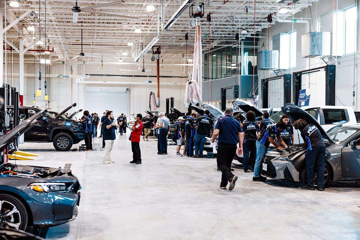 People work on cars in a spacious, well-lit automotive repair shop. Several vehicles have their hoods open, and technicians in uniforms are performing maintenance tasks as part of an Auto Tech Competition. Some people observe the process closely and take notes.