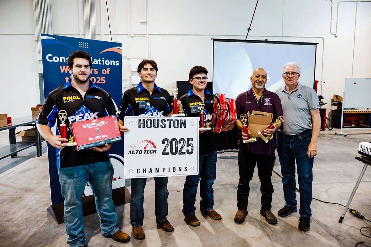 Four young men hold trophies and a “Houston 2025 Champions” sign, standing with two older men in a workshop. A banner behind them reads, “Congratulations Winners of 2025 Auto Tech Competition.” All are smiling and wearing casual attire.
