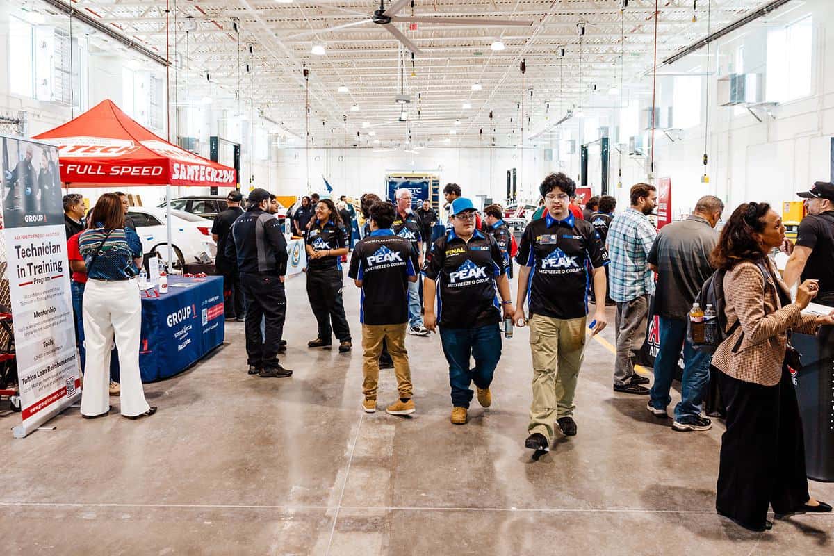 People attend an automotive training event in a large, bright garage, where the excitement of an Auto Tech Competition fills the air. Attendees interact at tables—some in uniforms engaged in technical discussions, while others observe or socialize. Cars and banners are visible in the background, adding to the dynamic atmosphere.