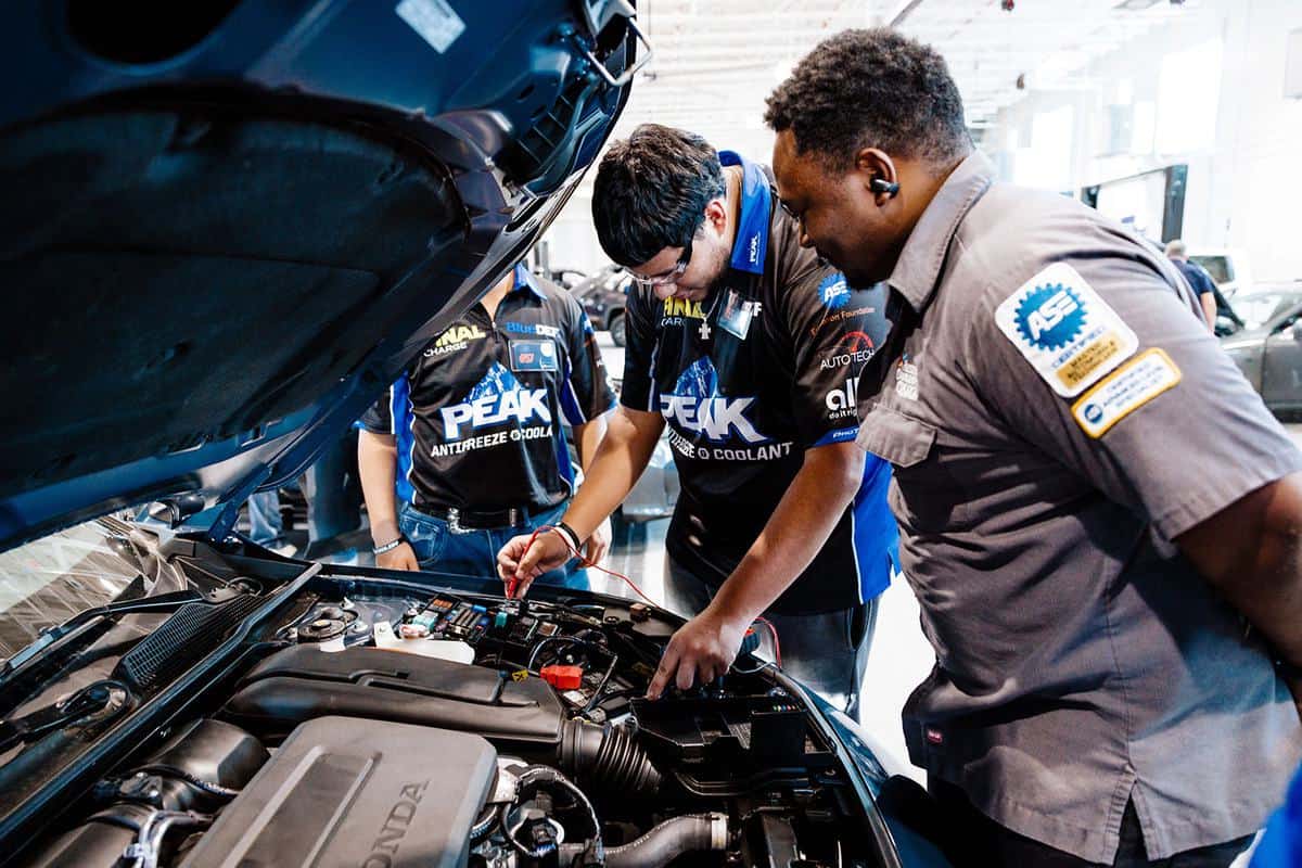 Two technicians examine a car engine with the hood open; one uses testing equipment while the other observes. Both wear uniforms with automotive logos and patches, showcasing their skills as they participate in an Auto Tech Competition, working in a well-lit garage.