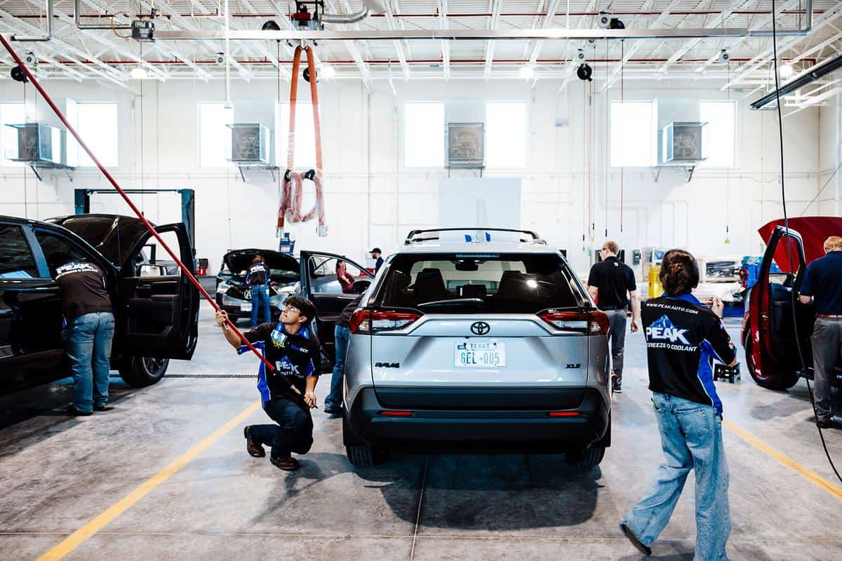 Several mechanics work on different vehicles inside a bright, spacious auto repair shop, creating an atmosphere reminiscent of an Auto Tech Competition. One person uses a tool overhead, while others inspect cars under the fluorescent lights.