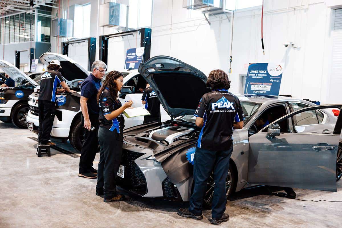 Several auto technicians in uniforms inspect cars with open hoods inside a well-lit repair garage, creating an atmosphere reminiscent of an Auto Tech Competition. One person writes on a clipboard, while others work on or examine the vehicles.